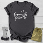 A dark charcoal gray t-shirt with 'Gymnastics Princess' printed in white script with a crown graphic, displayed on a white wood background alongside a pair of gymnastics bars and a gold drawstring bag.