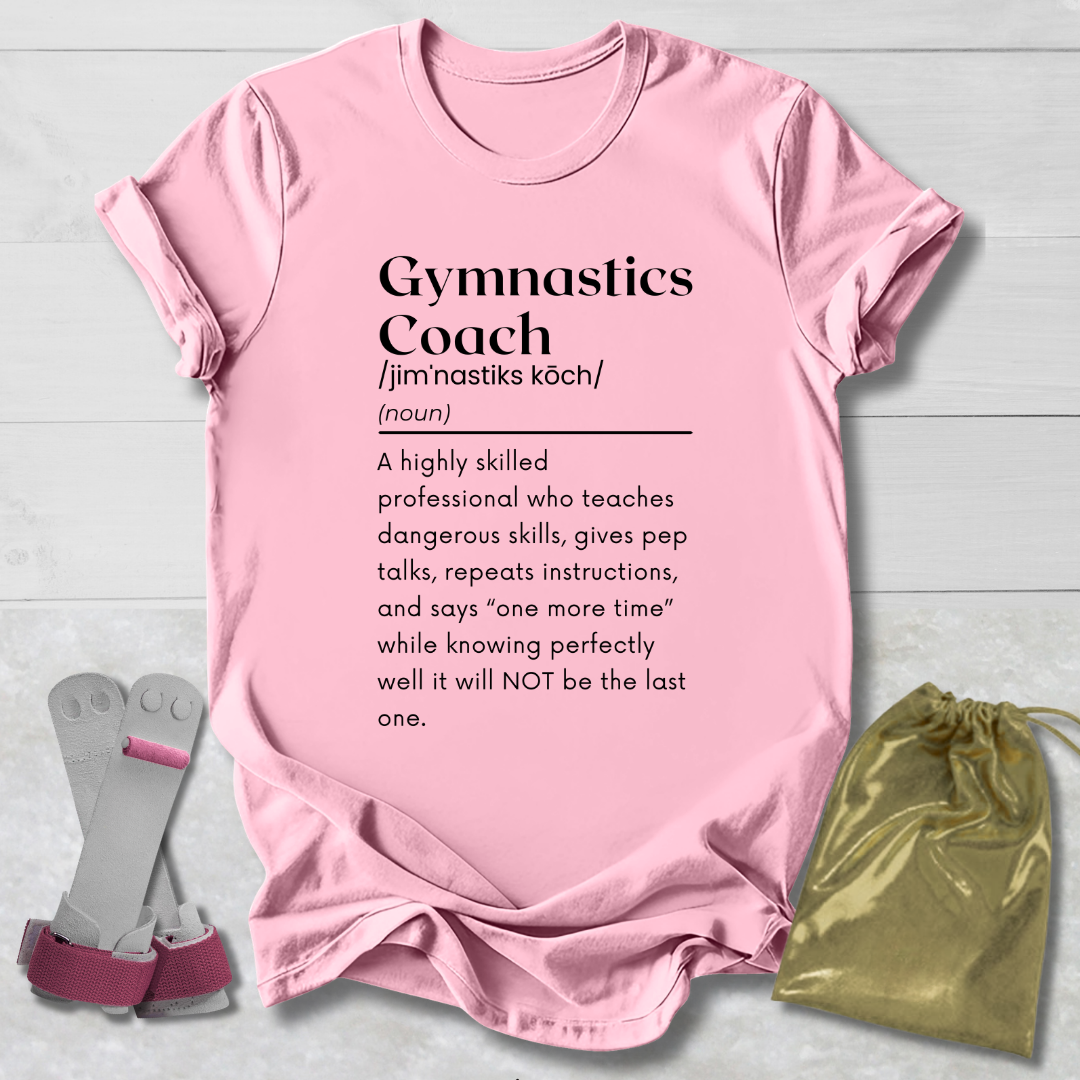 A light pink short-sleeve graphic T-shirt laid flat on a light wood background, with a gymnastics coach definition printed in black text. A pair of pink and gray gymnastics bars is placed to the left, and a small metallic gold drawstring pouch is to the right.