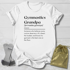 White  graphic t-shirt laid flat on a light wood background, featuring a black printed dictionary-style definition of 'Gymnastics Grandpa.' A pair of gymnastics trampoline bars and a gold drawstring pouch are placed beside the shirt as styling props.