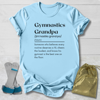 A light blue graphic t-shirt laid flat on a light wood background, featuring a black printed dictionary-style definition of 'Gymnastics Grandpa.' A pair of gymnastics trampoline bars and a gold drawstring pouch are placed beside the shirt as styling props.