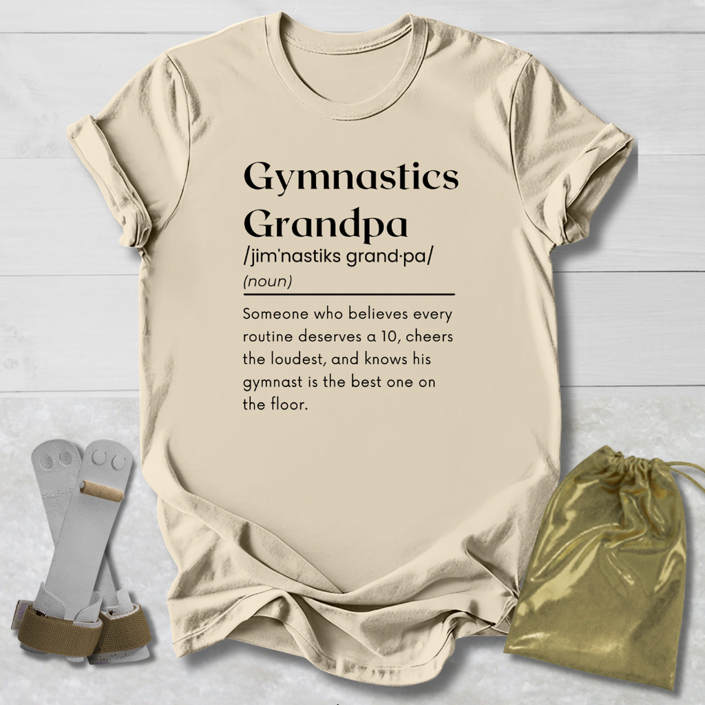 A beige graphic T-shirt laid flat on a light wood background, printed with the word 'Gymnastics' and a dictionary-style definition for 'Grandpa.' Two gray foam roller-style accessories and a small metallic gold drawstring pouch are placed beside the shirt as styling props.