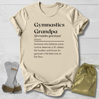 A beige graphic T-shirt laid flat on a light wood background, printed with the word 'Gymnastics' and a dictionary-style definition for 'Grandpa.' Two gray foam roller-style accessories and a small metallic gold drawstring pouch are placed beside the shirt as styling props.