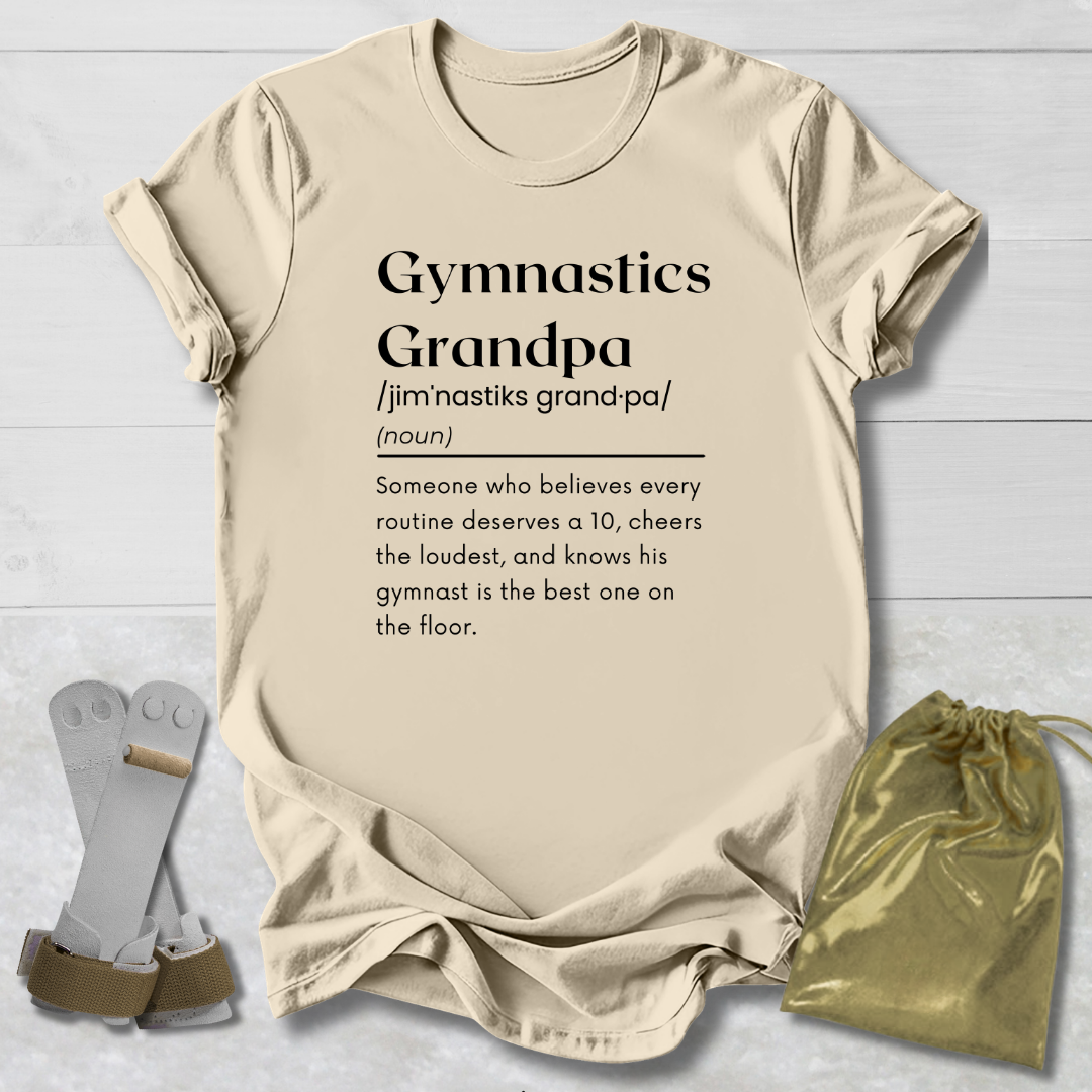 A beige graphic T-shirt laid flat on a light wood background, printed with the word 'Gymnastics' and a dictionary-style definition for 'Grandpa.' Two gray foam roller-style accessories and a small metallic gold drawstring pouch are placed beside the shirt as styling props.