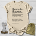 A beige graphic t-shirt laid flat on a light wood background, featuring a black printed definition of 'Gymnastics Grandma'. The shirt is styled with a pair of silver and gold gymnastics straps on the left and a small gold drawstring pouch on the right.