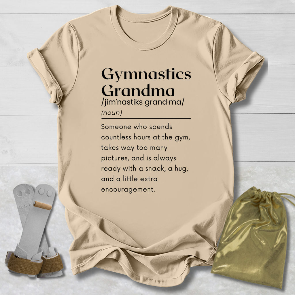 A beige graphic t-shirt laid flat on a light wood background, featuring a black printed dictionary-style definition of 'Gymnastics Grandma'. A pair of silver and tan gymnastics straps and a shiny gold drawstring pouch are styled around the shirt.