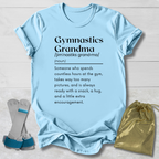 A light blue graphic t-shirt laid flat on a white wood background, featuring a black printed dictionary-style definition of 'Gymnastics Grandma.' A pair of gray and blue gymnastics bars sits to the left, and a shiny gold drawstring pouch is to the right.