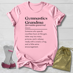 A light pink short-sleeve graphic T-shirt laid flat on a light gray wood background, with a gymnastics-themed dictionary definition printed on the front. A pair of pink-and-white gymnastics bars and a shiny gold drawstring pouch are placed beside the shirt as styling props.