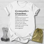 white short-sleeve graphic T-shirt laid flat on a light gray wood background, with a gymnastics-themed dictionary definition printed on the front. A pair of pink-and-white gymnastics bars and a shiny gold drawstring pouch are placed beside the shirt as styling props.