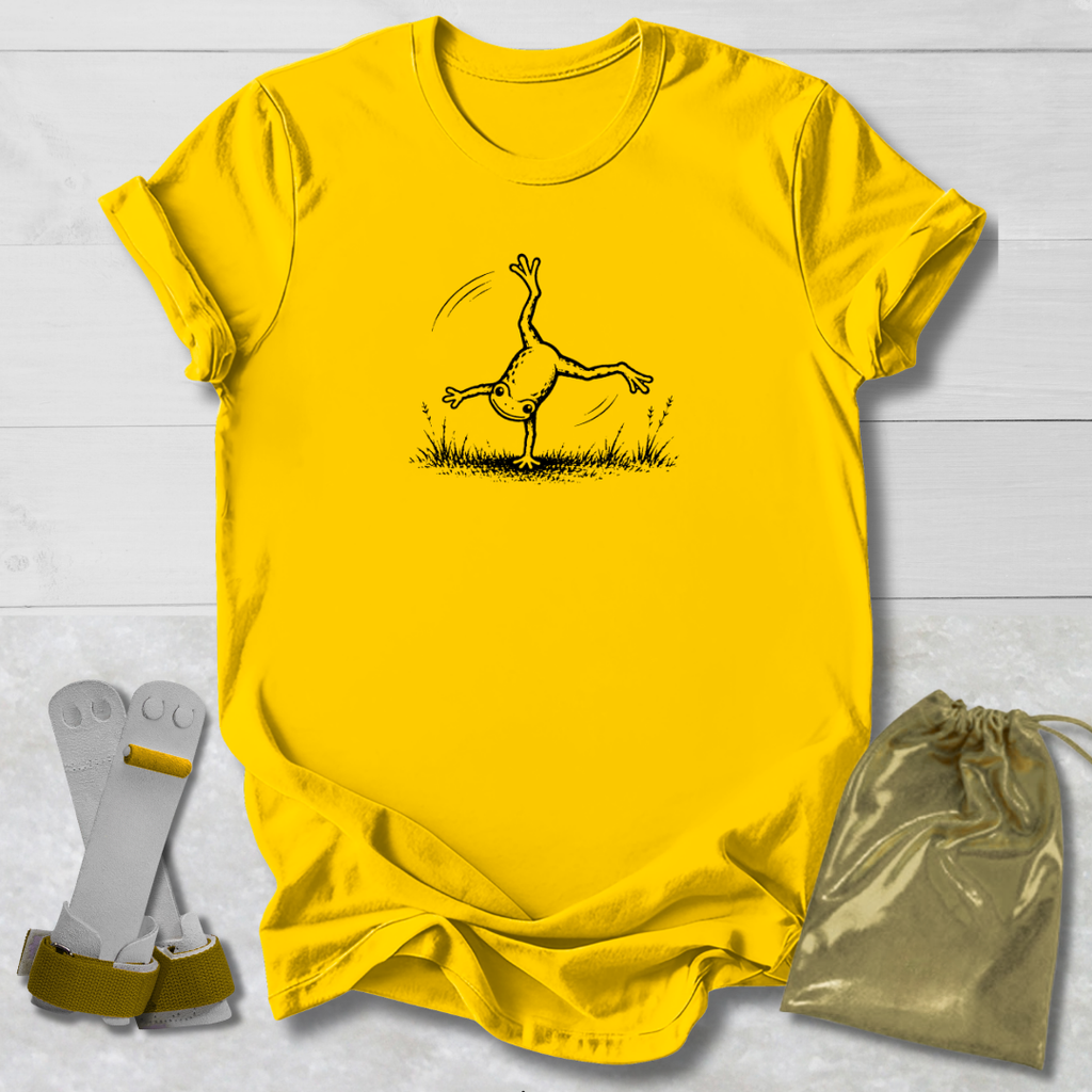 Yellow t-shirt with a black line art graphic of a frog doing a handstand, displayed on a white wood background alongside a gold drawstring bag and a pair of white and green wristbands.