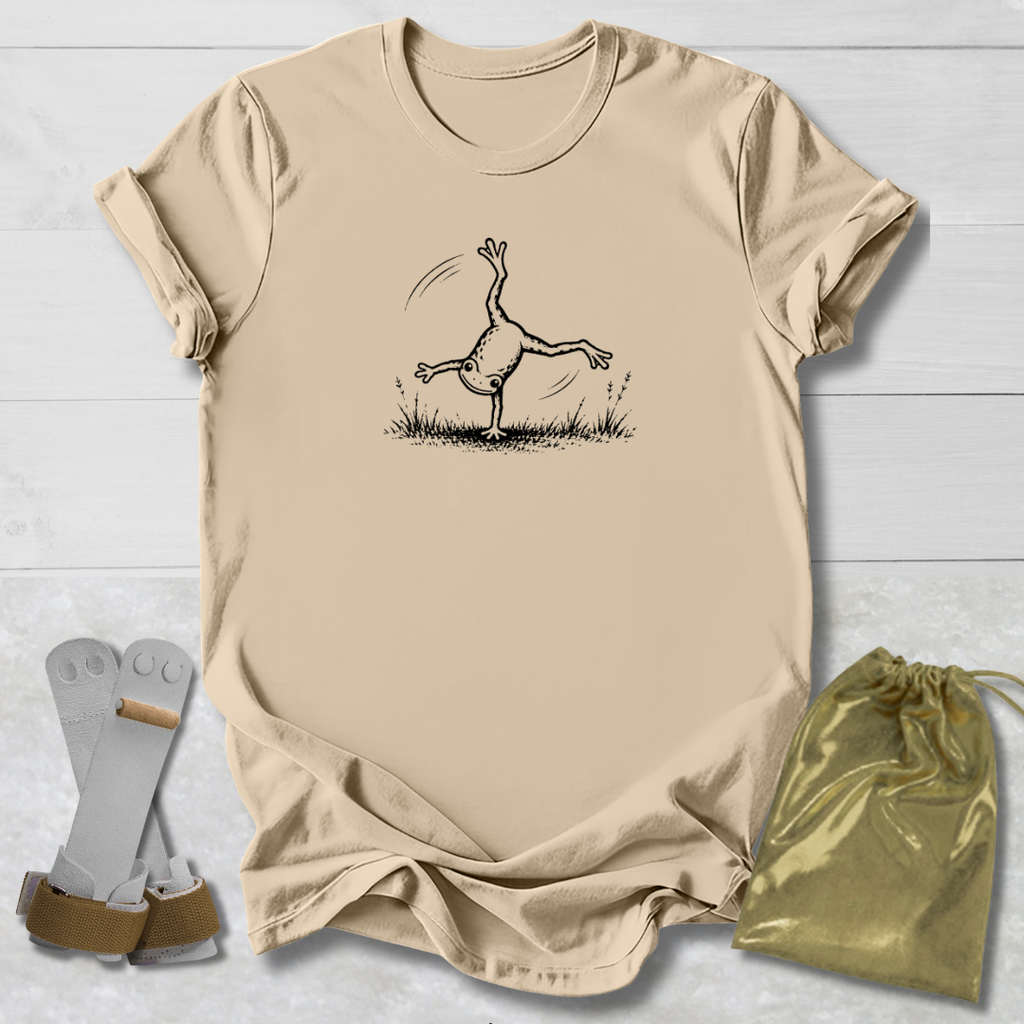 Beige t-shirt with a black line art graphic of a frog doing a handstand, displayed on a white wood background alongside a gold drawstring bag and a pair of white and green wristbands.