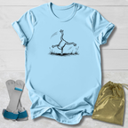 A light blue short-sleeve T-shirt laid flat on a white wood background, featuring a black line-art graphic of a frog balancing on one hand. A pair of gray and blue frog socks and a small gold drawstring pouch are placed beside the shirt as styling props.