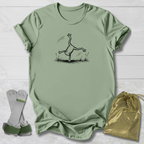 An olive green t-shirt with a black line art graphic of a frog doing a handstand, displayed on a white wood background alongside a gold drawstring bag and a pair of white and green wristbands.