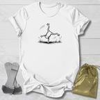 White t-shirt with a black line art graphic of a frog doing a handstand, displayed on a white wood background alongside a gold drawstring bag and a pair of white and green wristbands.