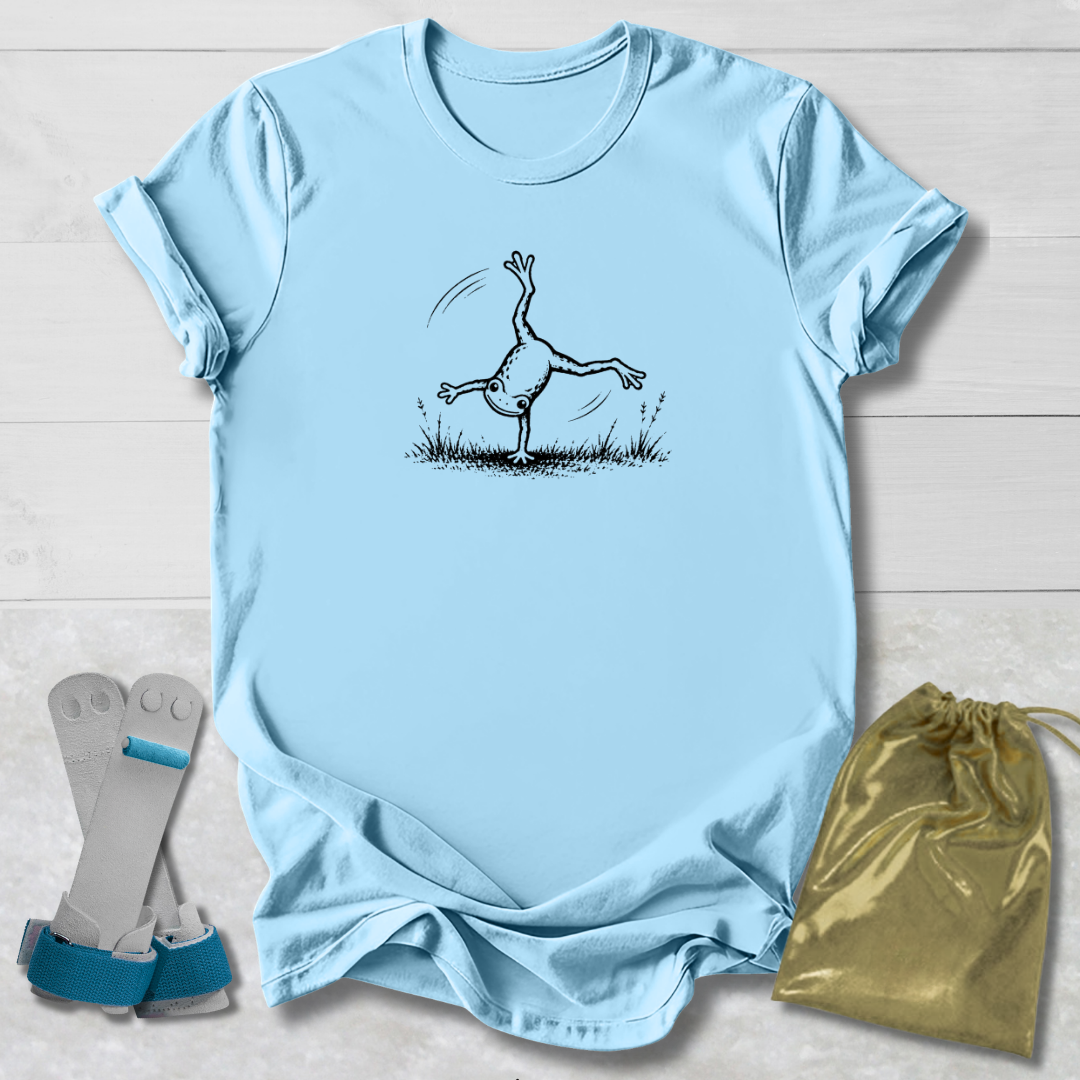 Blue t-shirt with a black line art graphic of a frog doing a handstand, displayed on a white wood background alongside a gold drawstring bag and a pair of white and green wristbands.