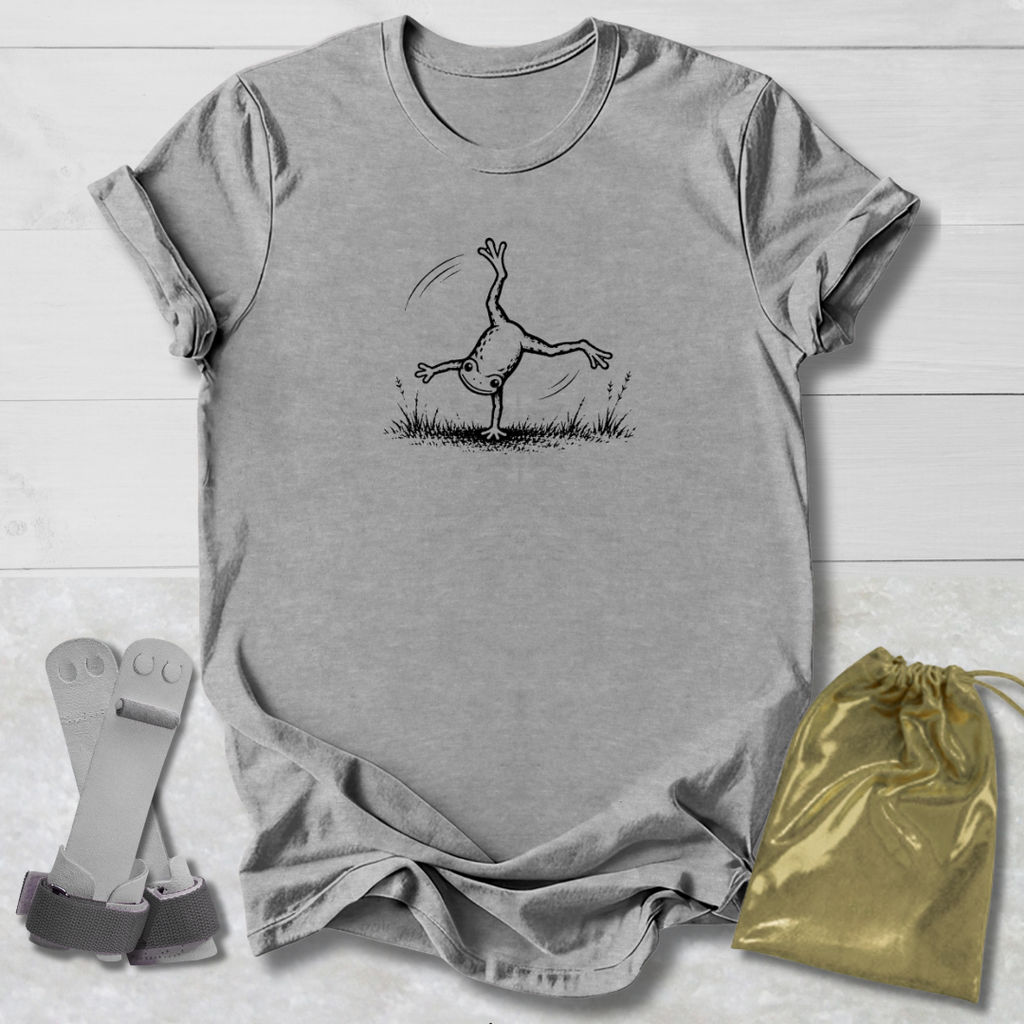 Gray t-shirt with a black line art graphic of a frog doing a handstand, displayed on a white wood background alongside a gold drawstring bag and a pair of white and green wristbands.