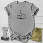 Gray t-shirt with a black line art graphic of a frog doing a handstand, displayed on a white wood background alongside a gold drawstring bag and a pair of white and green wristbands.