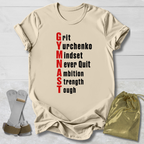 A beige graphic T-shirt laid flat on a light wood background, with rolled sleeves and a printed motivational gymnastics phrase in red and black text. A pair of gymnastics straps and a shiny gold drawstring pouch are placed beside the shirt as styling props.