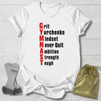 A white short-sleeve graphic T-shirt laid flat on a light wood background, with the phrase 'Grit Gymnchenko mindset never quit ambition strength toughness' printed vertically in red and black. A gold drawstring pouch and gray gymnastics straps are placed beside the shirt as styling props.