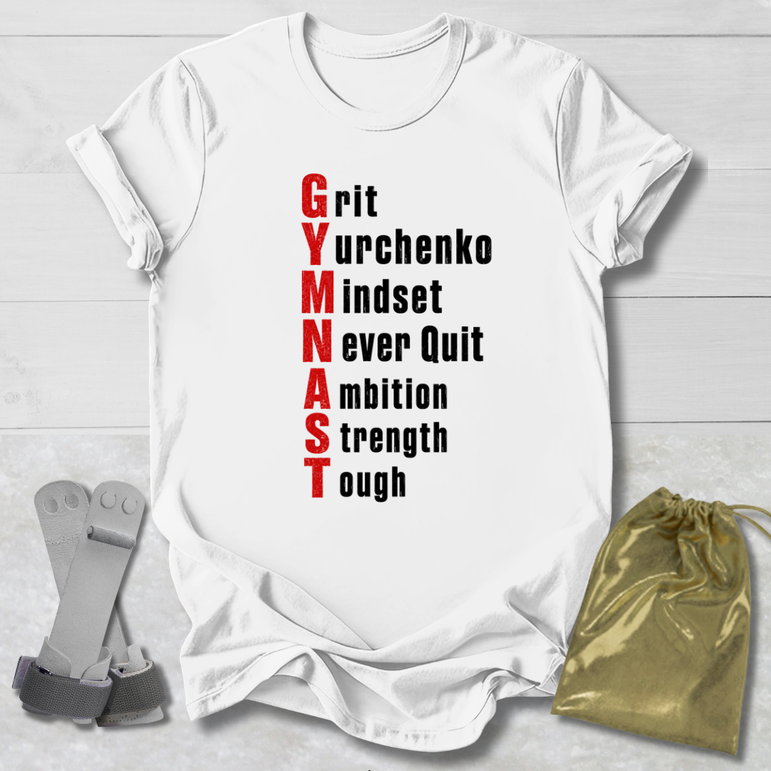 A white short-sleeve graphic T-shirt laid flat on a light wood background, with the phrase 'Grit Gymnchenko mindset never quit ambition strength toughness' printed vertically in red and black. A gold drawstring pouch and gray gymnastics straps are placed beside the shirt as styling props.