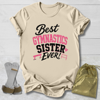 Beige  graphic T-shirt laid flat on a white wood background, printed with the phrase 'Best Gymnastics Sister Ever!' in black and pink lettering. A gold drawstring bag and a pair of gray gymnastics straps are placed beside the shirt as styling props.