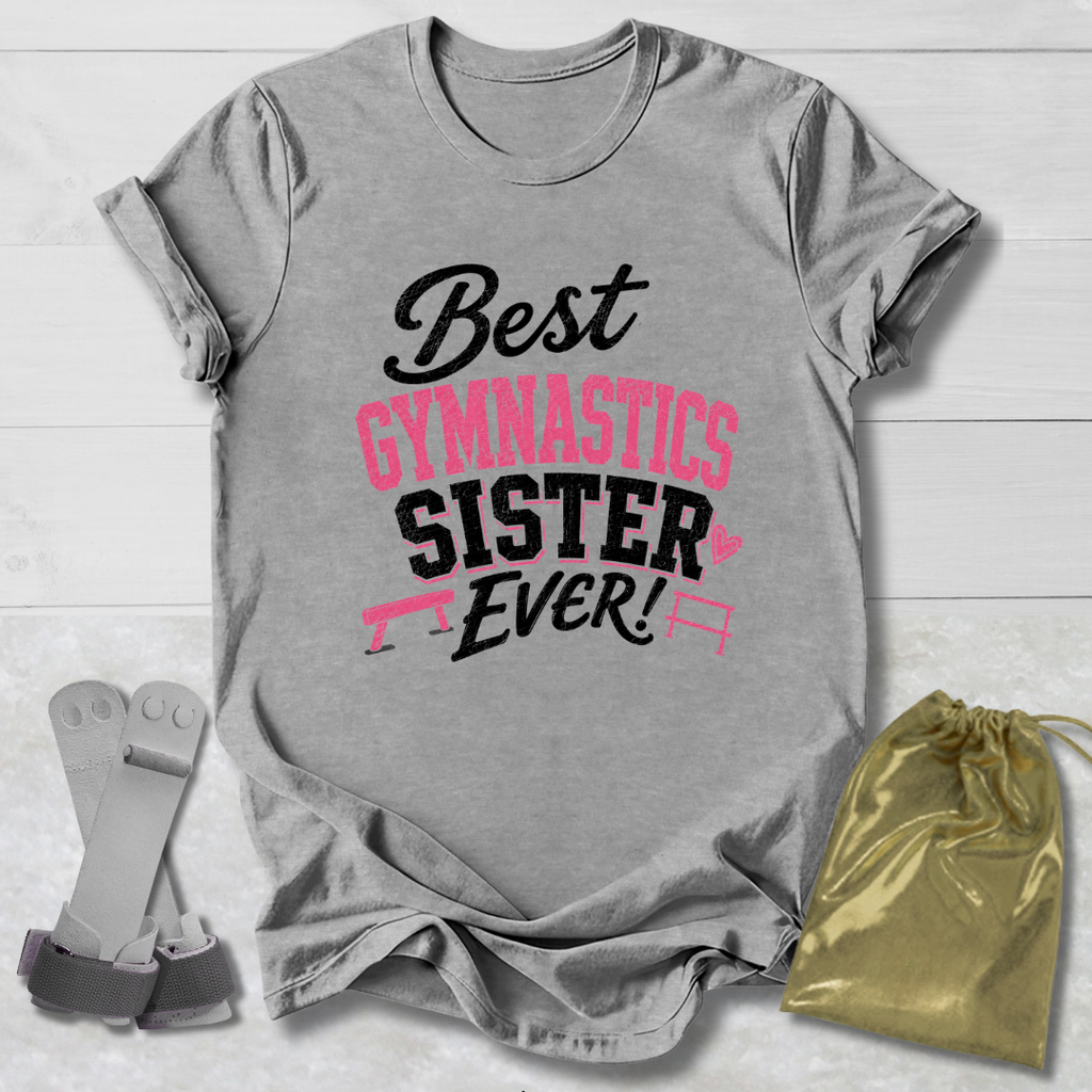A heather gray graphic T-shirt laid flat on a white wood background, printed with the phrase 'Best Gymnastics Sister Ever!' in black and pink lettering. A gold drawstring bag and a pair of gray gymnastics straps are placed beside the shirt as styling props.