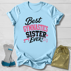 A light blue short-sleeve T-shirt laid flat on a white wood background, printed with the phrase 'Best Gymnastics Sister Ever!' in black and pink lettering with small gymnastics bar graphics. A pair of gymnastics shoes and a gold drawstring bag are placed beside the shirt as styling props.