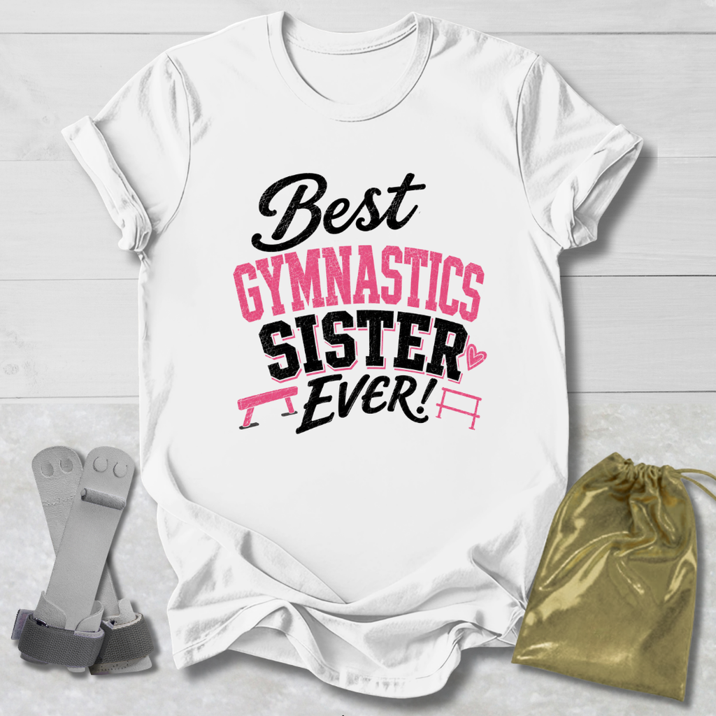 A white short-sleeve T-shirt laid flat on a light wood background, printed with the phrase 'Best Gymnastics Sister Ever!' in black and pink lettering with small gymnastics bar graphics. A gold drawstring pouch and a pair of gray straps are placed beside the shirt as styling props.