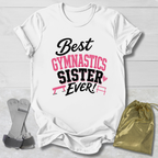 A white short-sleeve T-shirt laid flat on a light wood background, printed with the phrase 'Best Gymnastics Sister Ever!' in black and pink lettering with small gymnastics bar graphics. A gold drawstring pouch and a pair of gray straps are placed beside the shirt as styling props.