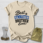 Beige short-sleeve T-shirt laid flat on a light wood background, printed with the phrase 'Best GYMNASTICS BROTHER Ever!' in black and blue lettering. A small black star appears between 'Best' and 'GYMNASTICS.