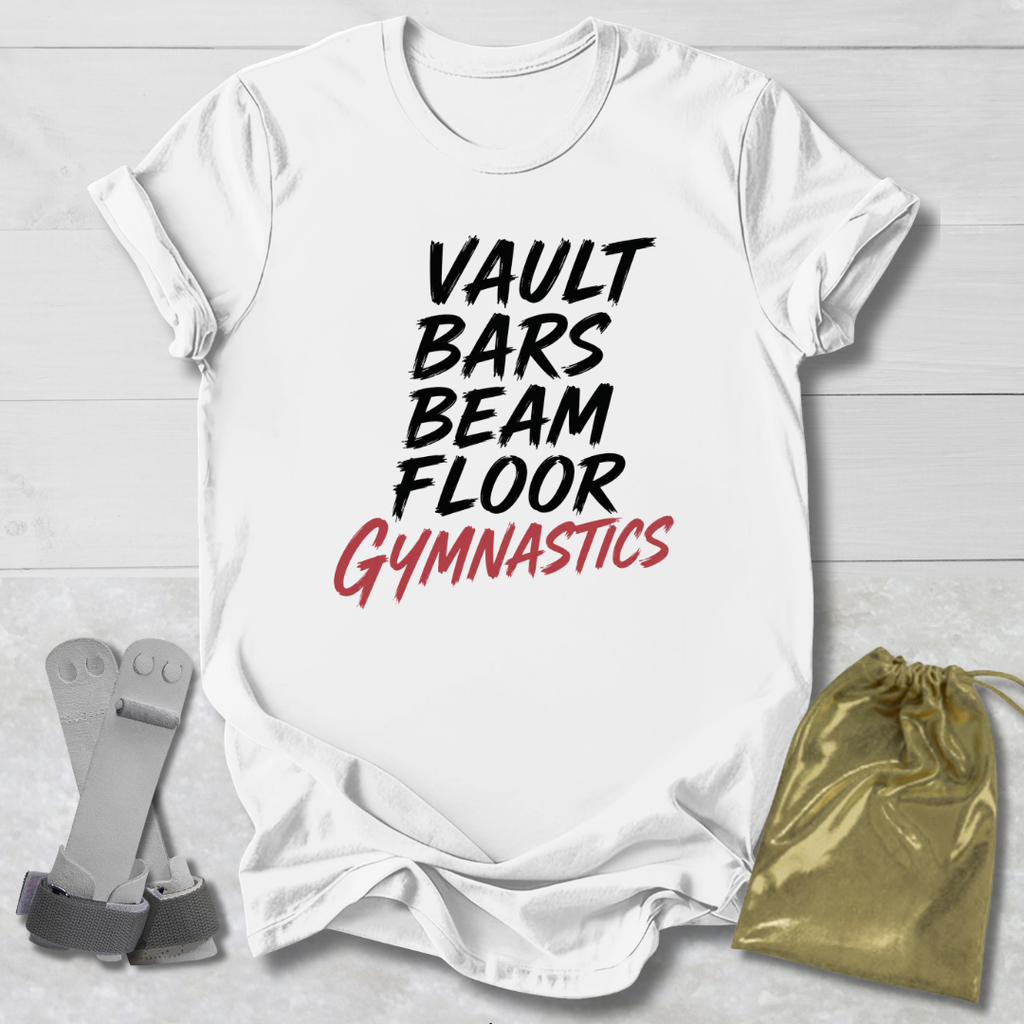 A white graphic T-shirt laid flat on a light wood background, with the text 'VAULT BARS BEAM FLOOR GYMNASTICS' printed on the front in black and red. A gold drawstring bag and gymnastics straps are placed beside the shirt as styling props.