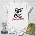 A white graphic T-shirt laid flat on a light wood background, with the text 'VAULT BARS BEAM FLOOR GYMNASTICS' printed on the front in black and red. A gold drawstring bag and gymnastics straps are placed beside the shirt as styling props.