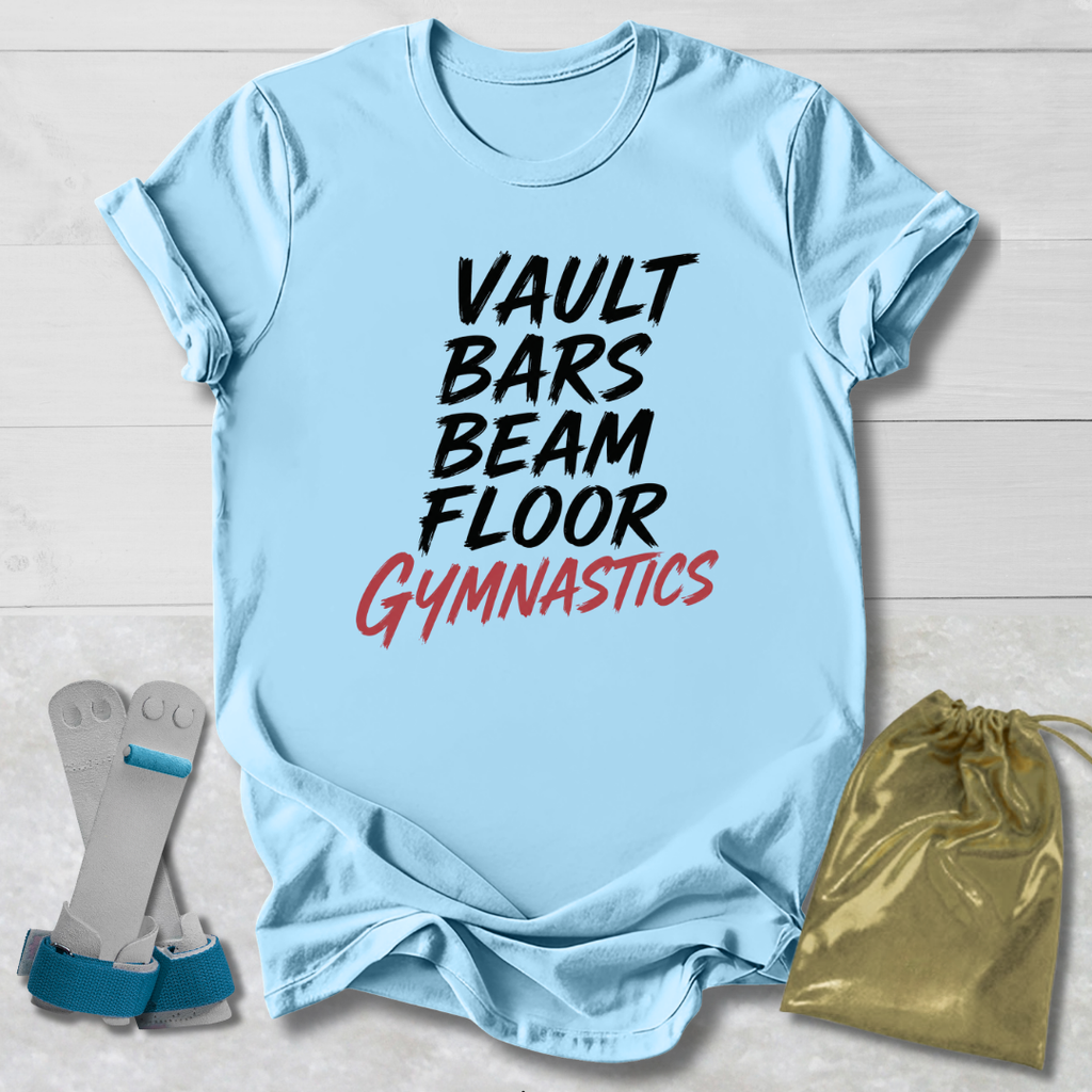 A light blue graphic T-shirt laid flat on a white wood background, with the words 'VAULT BARS BEAM FLOOR GYMNASTICS' printed on the front in black and red. A pair of gymnastics shoes and a gold drawstring bag are placed beside the shirt as styling props.