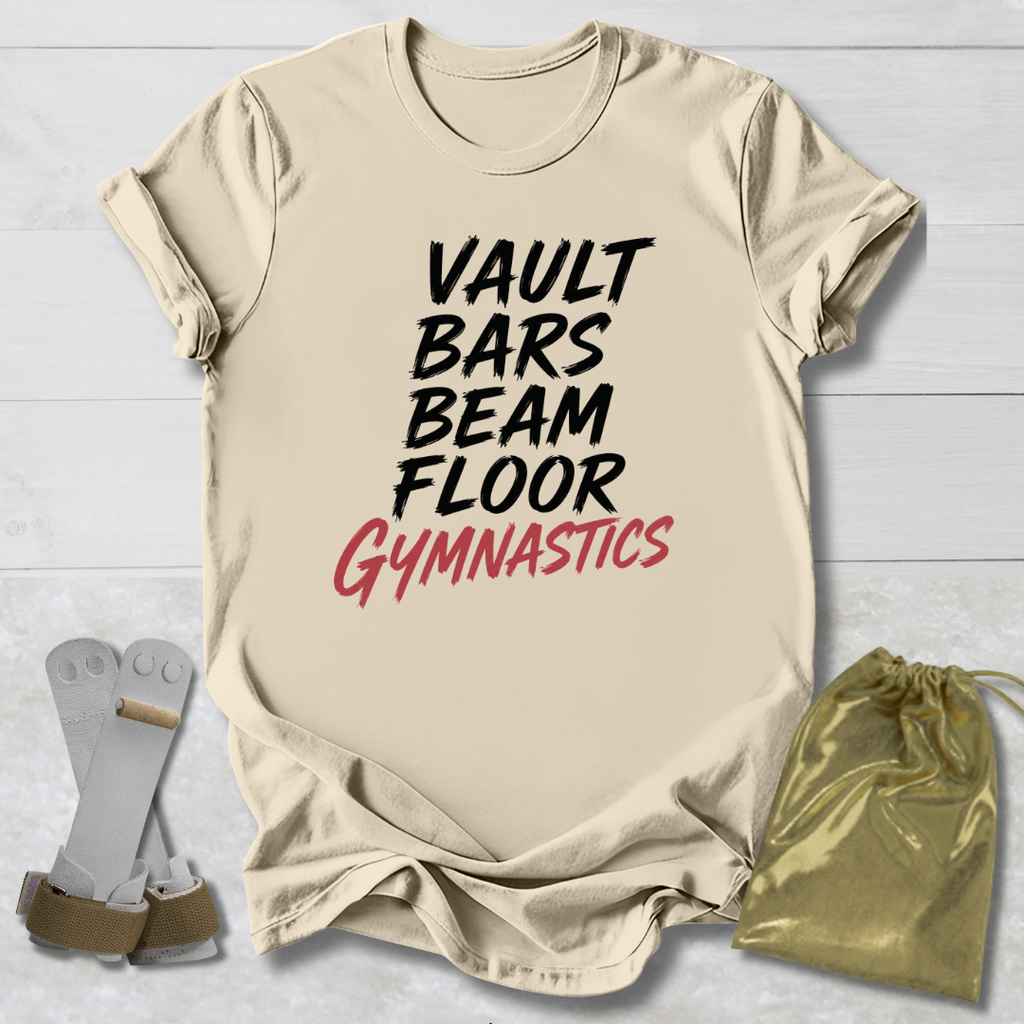 A beige graphic T-shirt laid flat on a light wood background, with the words 'VAULT BARS BEAM FLOOR GYMNASTICS' printed on the front in black and red. A pair of gymnastics bars and a shiny gold drawstring bag are placed beside the shirt as styling props.
