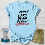 A light blue graphic T-shirt laid flat on a white wood background, with the words 'VAULT BARS BEAM FLOOR GYMNASTICS' printed on the front in black and red. A pair of gymnastics shoes and a gold drawstring bag are placed beside the shirt as styling props.