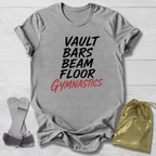 A heather gray graphic t-shirt laid flat on a white wood background, featuring the text 'VAULT BARS BEAM FLOOR GYMNASTICS' in black and red. A gold drawstring bag and gymnastics vaulting straps are placed beside the shirt.