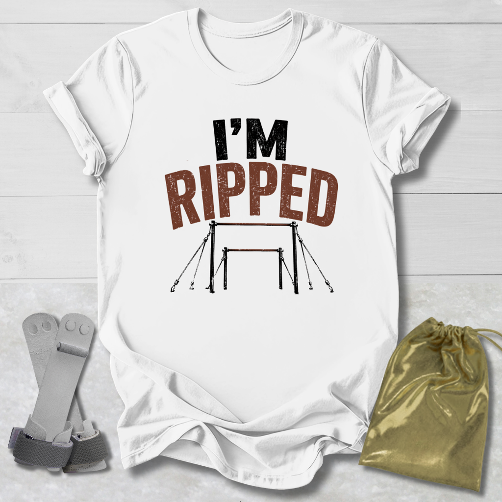 A white graphic t-shirt laid flat on a light wood background, featuring the text 'I'M RIPPED' with a gymnastics bar graphic. A gold drawstring bag and a pair of gray straps are placed beside the shirt as props.