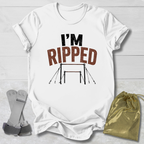 A white graphic t-shirt laid flat on a light wood background, featuring the text 'I'M RIPPED' with a gymnastics bar graphic. A gold drawstring bag and a pair of gray straps are placed beside the shirt as props.