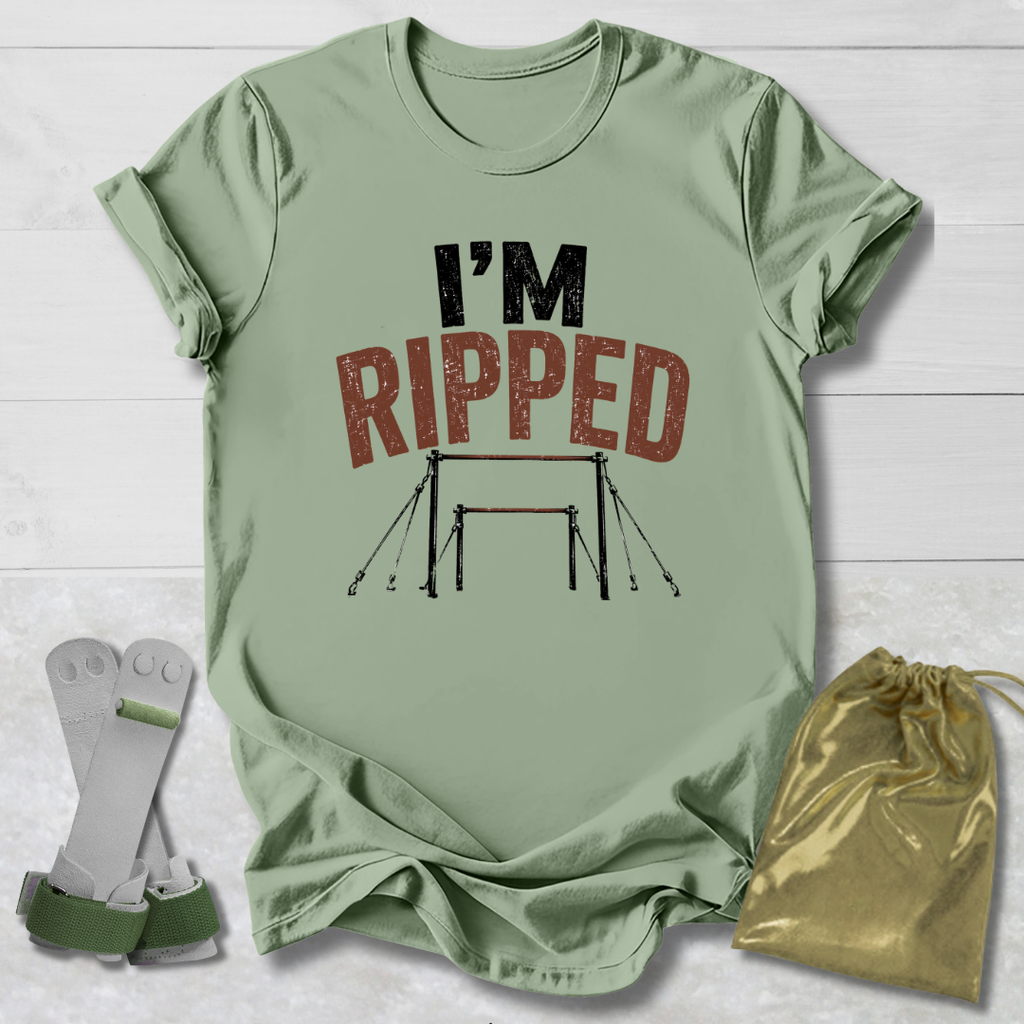A sage green graphic t-shirt laid flat on a white wood background, featuring the text 'I'M RIPPED' with a gymnastics bar graphic. A pair of white and green gymnastics straps and a gold drawstring bag are placed beside the shirt.