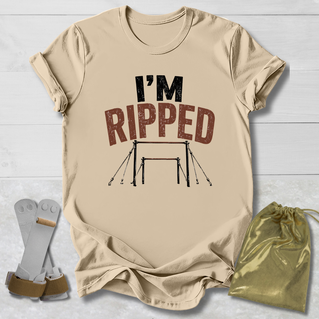 A beige graphic T-shirt laid flat on a white wood background, with the text 'I'M RIPPED' and a gymnastics bar graphic printed on the front. A pair of gymnastics straps and a small gold drawstring bag are placed beside the shirt as styling props.