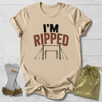 A beige graphic T-shirt laid flat on a white wood background, with the text 'I'M RIPPED' and a gymnastics bar graphic printed on the front. A pair of gymnastics straps and a small gold drawstring bag are placed beside the shirt as styling props.