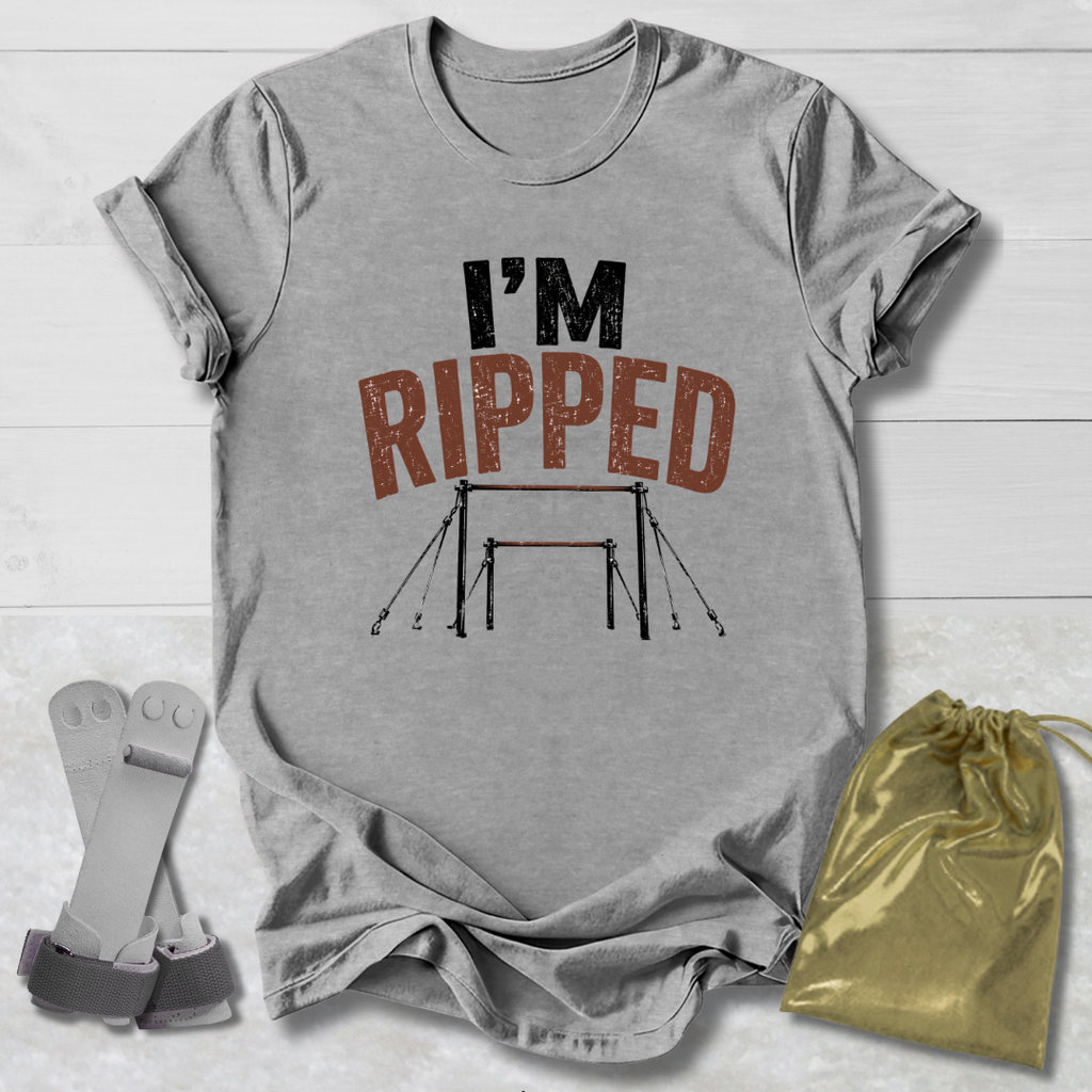 A heather gray graphic t-shirt laid flat on a white wood background, featuring the text 'I'M RIPPED' with a gymnastics bar graphic. A gold drawstring bag and a pair of gray straps are placed beside the shirt.