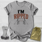A heather gray graphic t-shirt laid flat on a white wood background, featuring the text 'I'M RIPPED' with a gymnastics bar graphic. A gold drawstring bag and a pair of gray straps are placed beside the shirt.