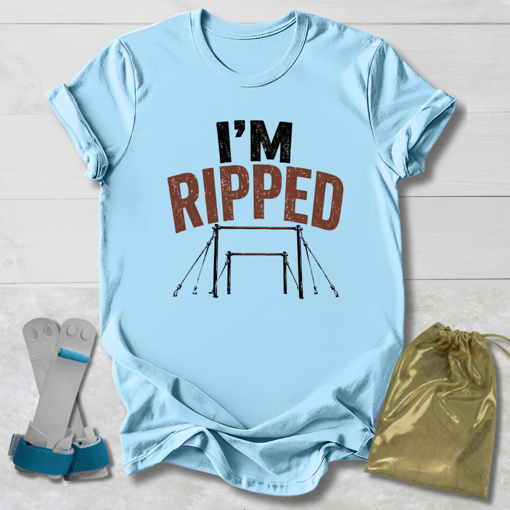 A light blue graphic T-shirt laid flat on a white wood background, with the words “I’M RIPPED” and a gymnastics bar graphic printed on the front. A pair of gymnastics straps and a gold drawstring bag are placed beside the shirt as styling props.