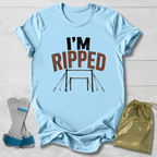 A light blue graphic T-shirt laid flat on a white wood background, with the words “I’M RIPPED” and a gymnastics bar graphic printed on the front. A pair of gymnastics straps and a gold drawstring bag are placed beside the shirt as styling props.