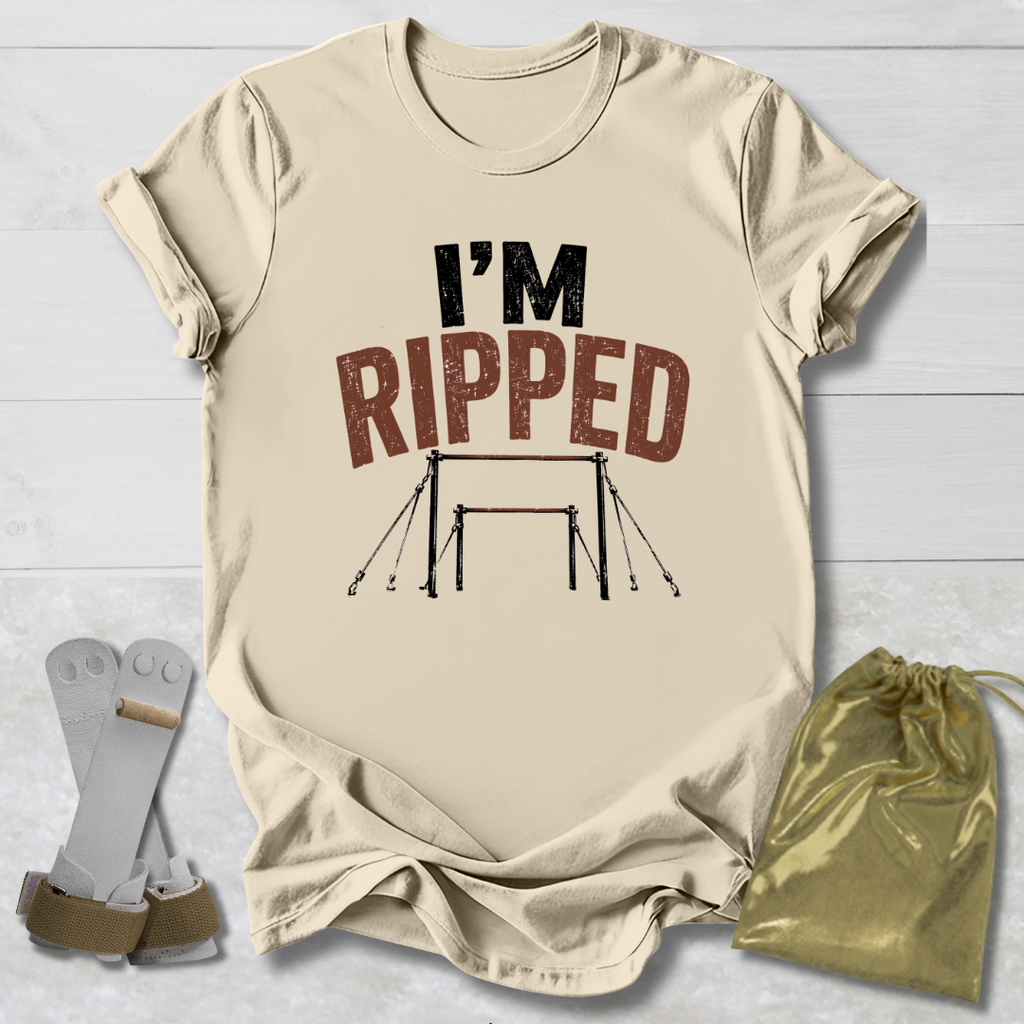 A beige graphic t-shirt laid flat on a white wood background, featuring the text 'I'M RIPPED' with a gymnastics bar graphic. A pair of gymnastics straps and a gold drawstring bag are placed beside the shirt.