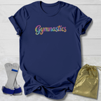 A navy blue short-sleeve t-shirt with the word 'Gymnastics' printed across the chest in a colorful glitter-style font. The shirt is displayed in a flat-lay arrangement on a white wood background, accompanied by a gold drawstring bag and a pair of gymnastics wrist wraps.