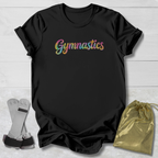 A black short-sleeve T-shirt laid flat on a light wood background, featuring the word 'Gymnastics' across the chest in a colorful glitter-style multicolor font. A pair of gymnastics bars and a small metallic gold drawstring bag are placed beside the shirt as styling props.