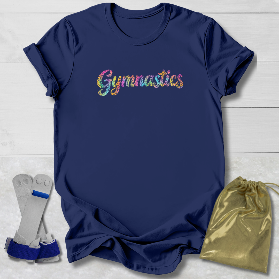 A navy blue short-sleeve t-shirt with the word 'Gymnastics' printed across the chest in a colorful glitter-style font. The shirt is displayed in a flat-lay arrangement on a white wood background, accompanied by a gold drawstring bag and a pair of gymnastics wrist wraps.