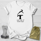 A white short-sleeve graphic T-shirt laid flat on a light wood background, featuring a black gymnast silhouette on a balance beam with the text 'just one of those days.' Gray diving boards and a gold drawstring pouch are placed beside the shirt as styling props.