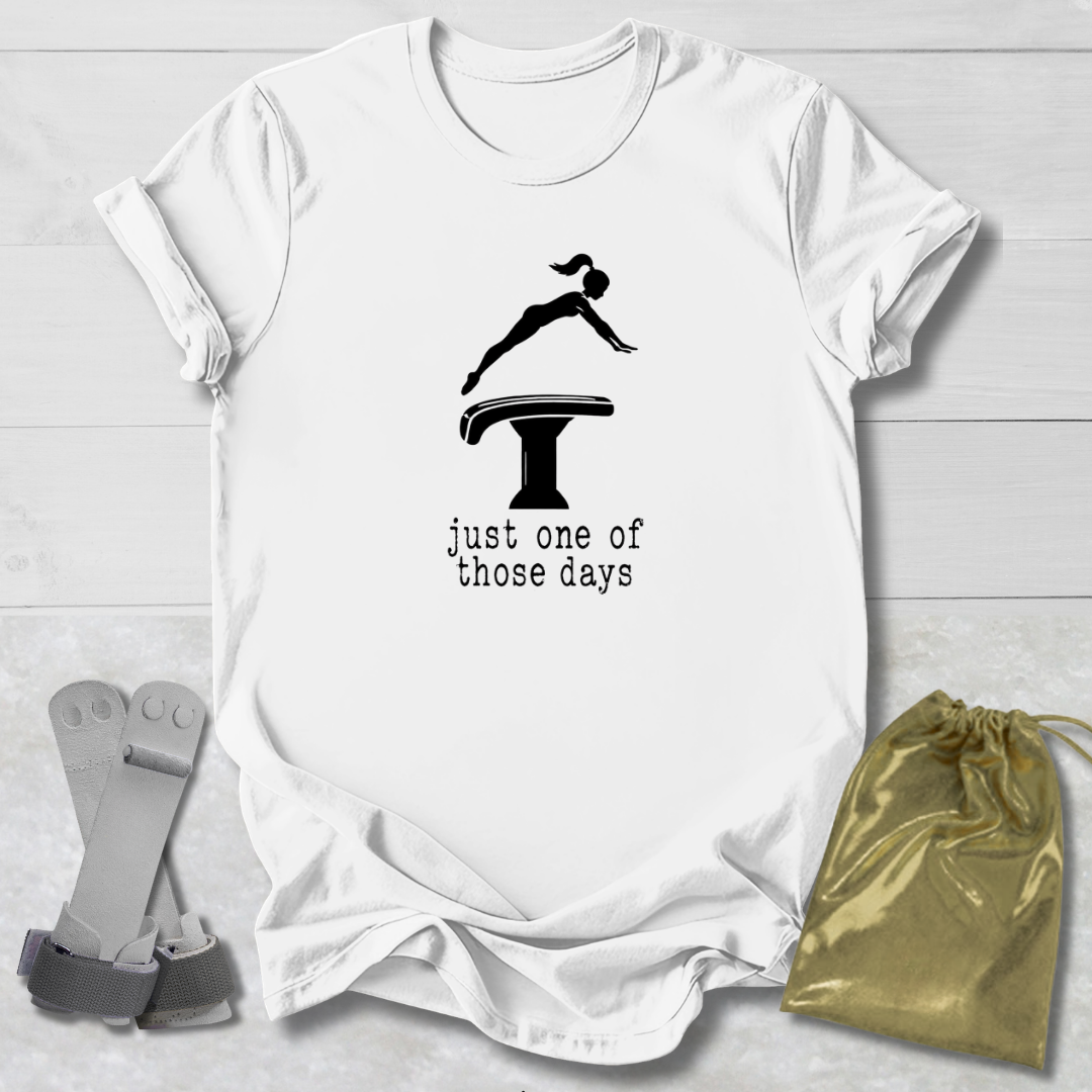 A white short-sleeve graphic T-shirt laid flat on a light wood background, featuring a black gymnast silhouette on a balance beam with the text 'just one of those days.' Gray diving boards and a gold drawstring pouch are placed beside the shirt as styling props.
